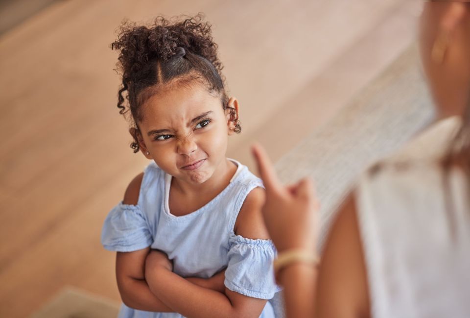 What is a positive behavior support plan? Why your child needs one. A young black girl in a light blue ruffled top is making a displeased face with furrowed brows while standing indoors. She is crossing her arms and pointing upwards with one finger, expressing disapproval or making a point during a discussion with an adult, whose presence is implied but not shown in the image. The child's curly hair is styled in small buns.