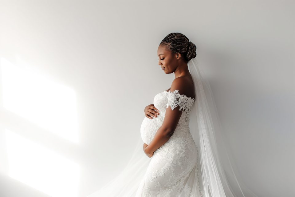 A glowing pregnant black bride in a lace wedding dress stands in profile, showing how to plan your wedding when pregnant with beauty and confidence.