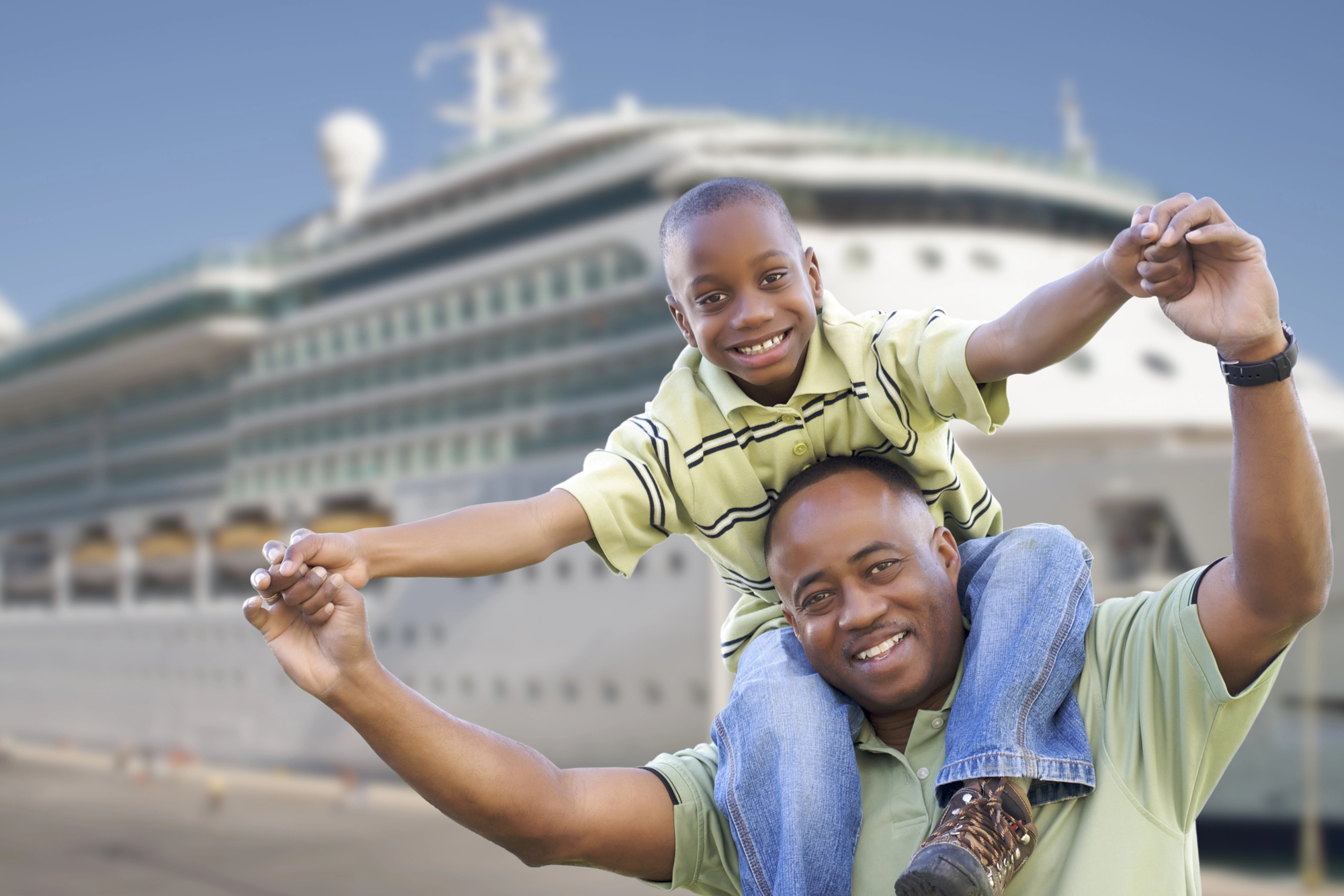 Luxury travel on a budget: finding deals on cruises An african american father with his son on his shoulder are standing in the foreground of a cruise ship for an article about a cruise ship on turquoise water for an article on luxury travel on a budget: finding deals on cruises