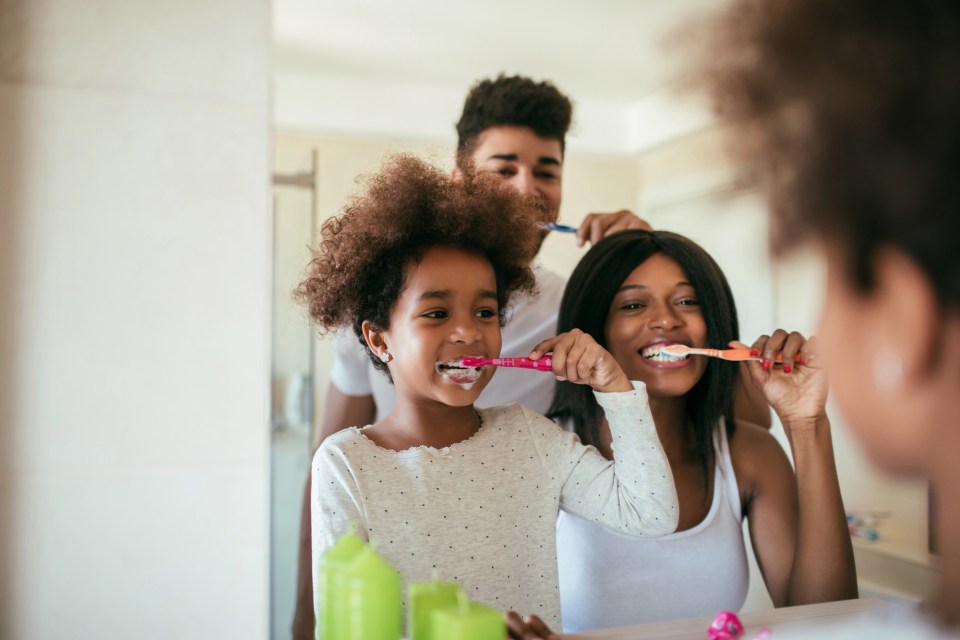 Caring for your family’s teeth: a parent’s guide An african american family brushes their teeth together in a mirror to care for their family's teeth as part of a parent's guide.