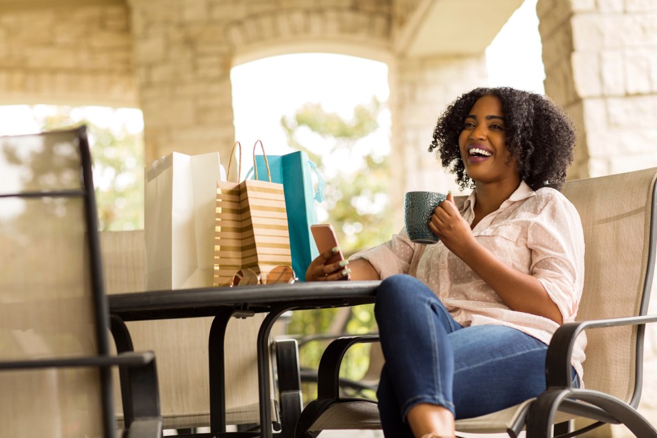 Gifts for loved ones who need a boost A joyful woman enjoying a coffee while shopping for gifts for loved ones. She is surrounded by shopping bags on a sunny day, symbolizing the happiness and satisfaction that comes from thoughtful gift-giving.