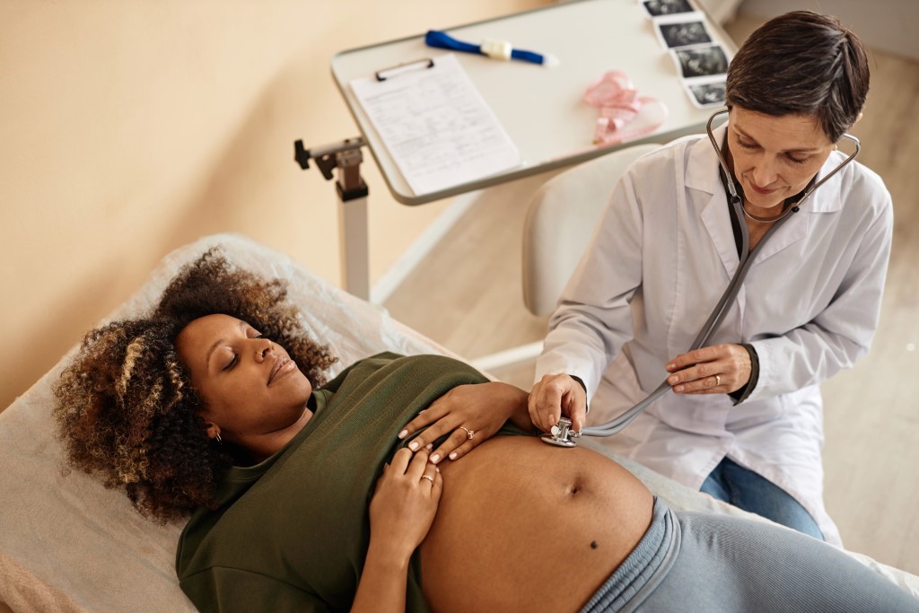 A prenatal care provider listens to the abdomen of a pregnant woman with a stethoscope during a routine pregnancy checkup in a clinical setting.