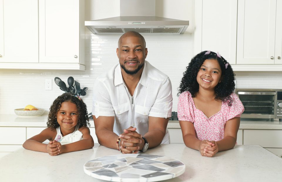 Dad jokes on deck: finesse mitchell’s best role yet Finesse mitchell leans on a white kitchen countertop between his daughters—eva kristian on his left and elle kate on his right—all three smiling warmly into the camera.