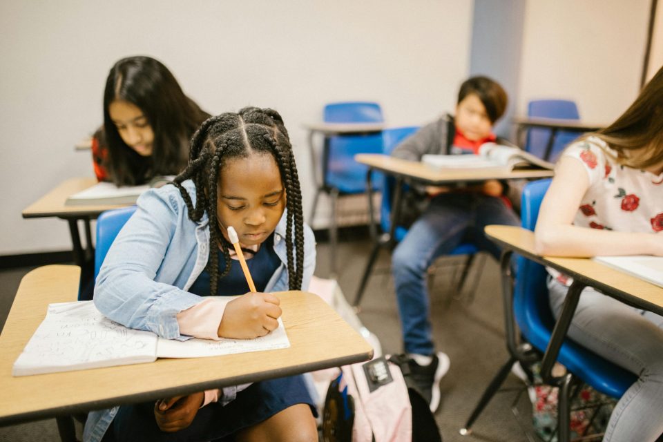 A young black girl writing in a workbook at her school desk, surrounded by classmates in a classroom setting—illustrating the importance of student safety and focused learning supported by school access control systems.