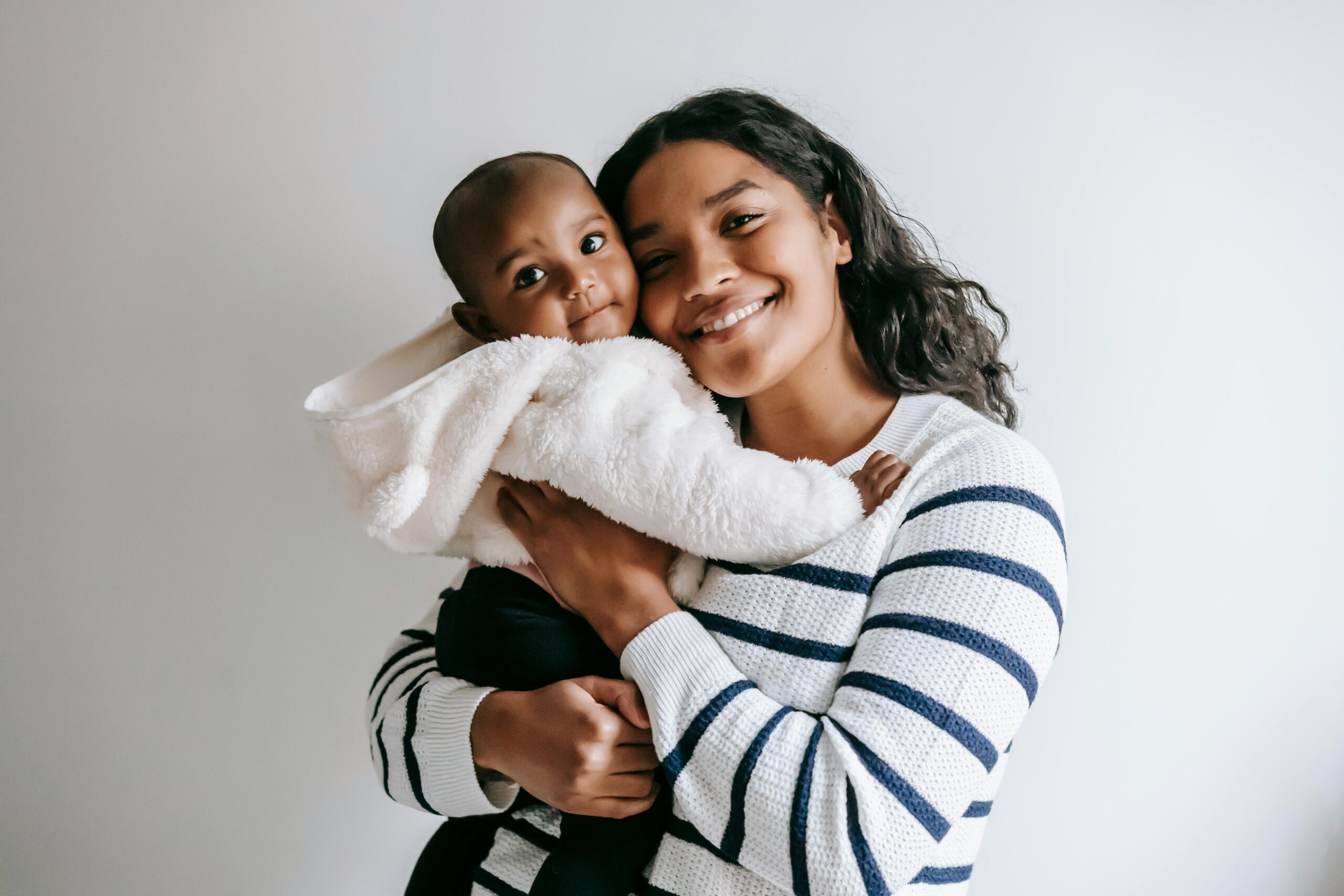 What you can do about being tired as a parent A tired black mother in a striped sweater smiles warmly while holding her baby wrapped in a soft white jacket against a plain light background.