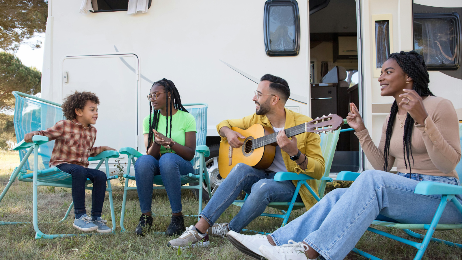 A family of four sits in folding lawn chairs outside a camper van; a young black boy, a black teenage girl, a father playing guitar, and a black mother smiling and singing along.