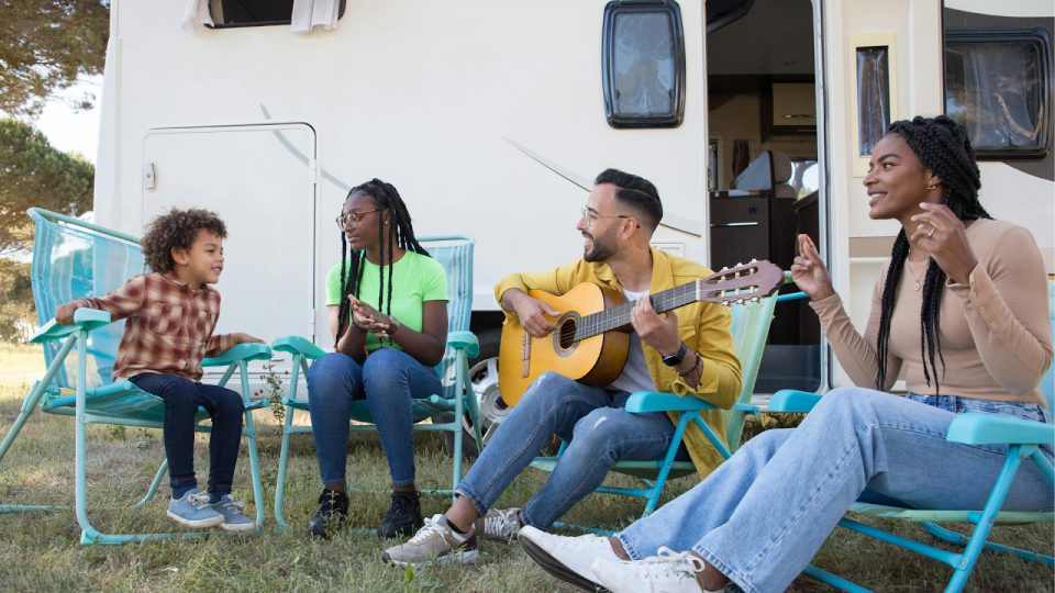 7 smart tips for traveling with kids this summer (without losing your mind) A family of four sits in folding lawn chairs outside a camper van; a young black boy, a black teenage girl, a father playing guitar, and a black mother smiling and singing along.