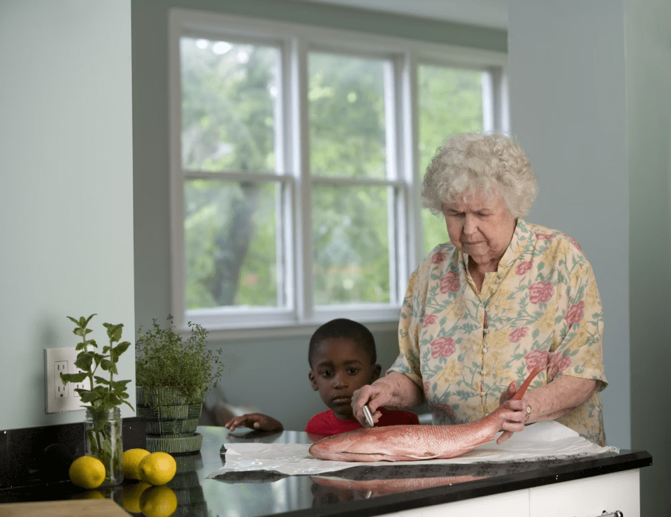 An elderly woman prepares a whole fish at the kitchen counter while a young black boy watches attentively, symbolizing intergenerational caregiving and early signs of when assisted care may be needed.