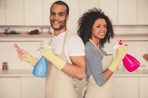 How to create a chore chart that actually works for busy black families Black couple smiling while holding cleaning spray bottles and wearing aprons—representing teamwork and shared chores in black families for an article about how to create a chore chart that actually works for busy black families
